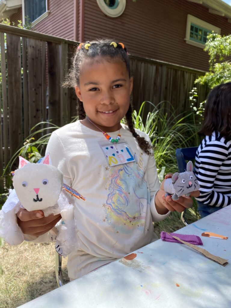 Young camper holds up two creative creature creations. On the left she is holding a white cat with pink nose and ears made out of felt and cotton balls. on the right she is holding a small mouse with rounded ears and drawn-on whiskers, made out of grey felt.
