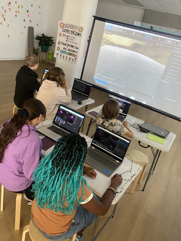 Five campers are seated at three tables working on laptops, facing a projector screen that is displaying a program used for virtual world design.