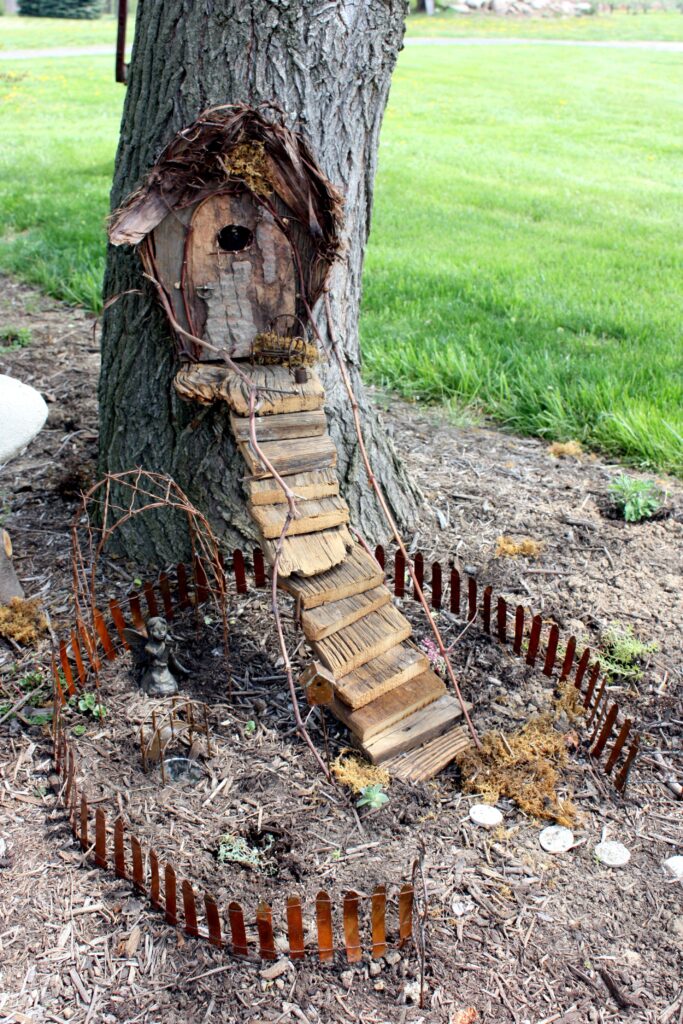 Handmade fairy house built into the base of a tree, featuring a rustic wooden ladder, miniature fencing, and tiny garden decorations.