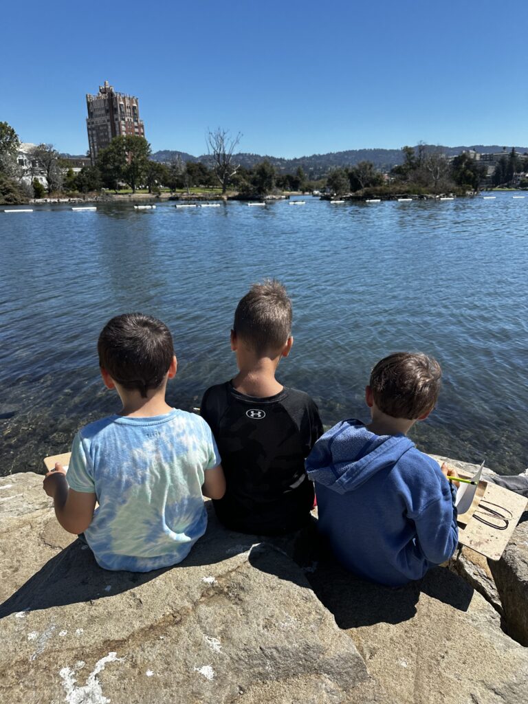 Three young camper sit on the rocky shore of Lake Merritt in Oakland, writing and drawing in the Design Journals with the beautiful lake behind them.
