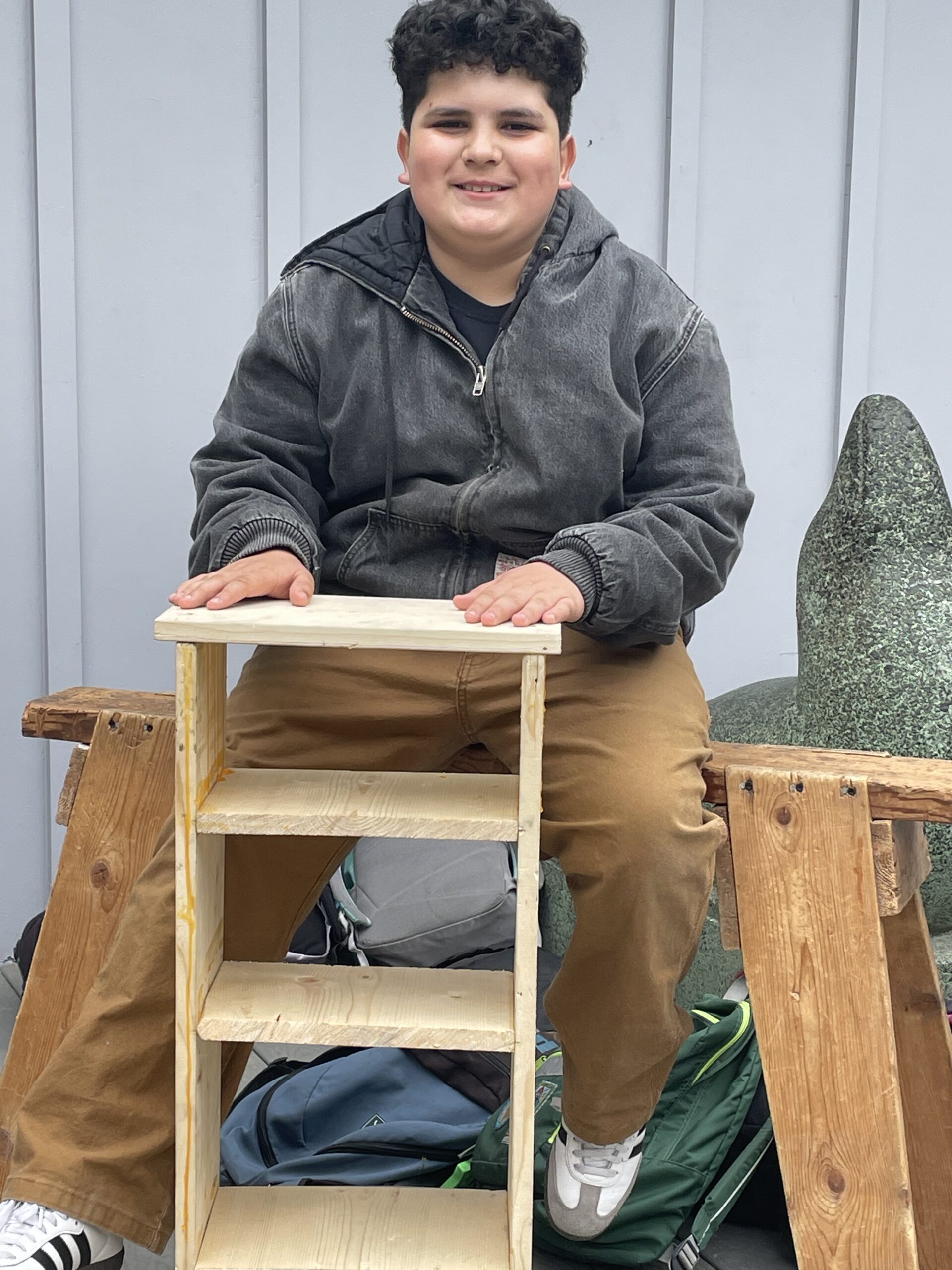 Smiling camper seated on a saw horse, posing with his bookshelf made in camp.