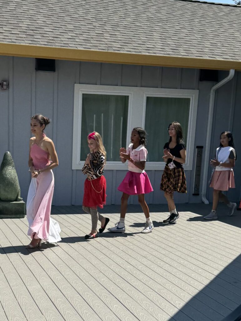 An action shot of five fashion design campers walking the runway in their finished pieces. The first camper is in a one shoulder dress with pink bodice and half pink half white floor length skirt. the second camper is in a cheetah print long sleeve top and red mid length skirt with a matching red bow in her hair. The third camper is in light pink polo shirt with a white collar and hot pink circle skirt. The forth camper is in a black t-shirt and yellow, black, and red plaid mid length skirt. The last camper is in a loose fitting white tank top and light pink mid length skirt.