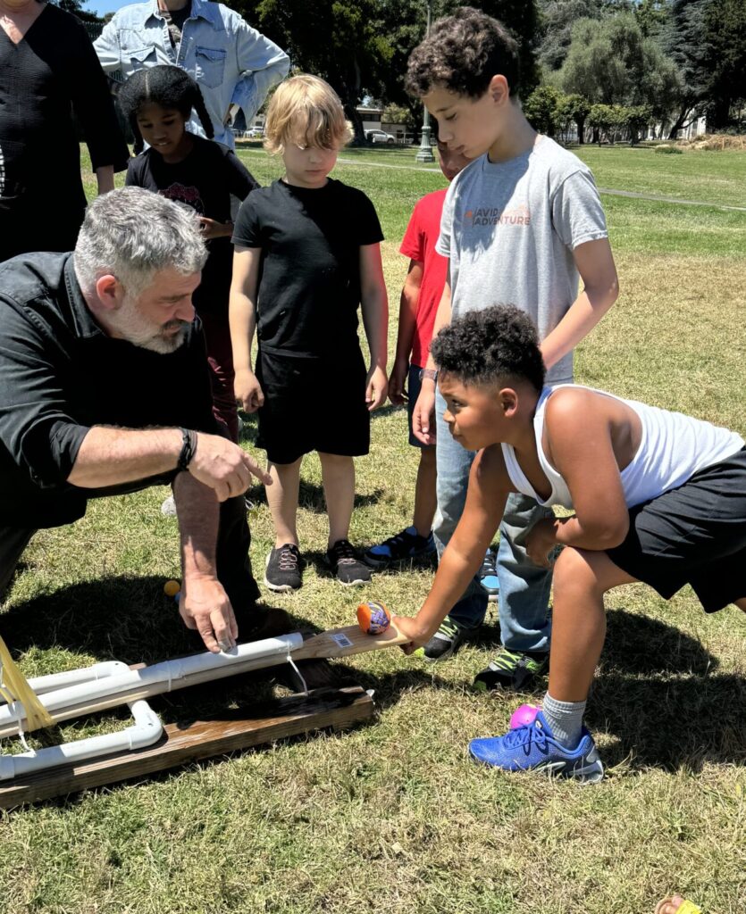 Three campers gather around a catapult trebuchet with their teacher outside on a lawn, ready to launch a ball.