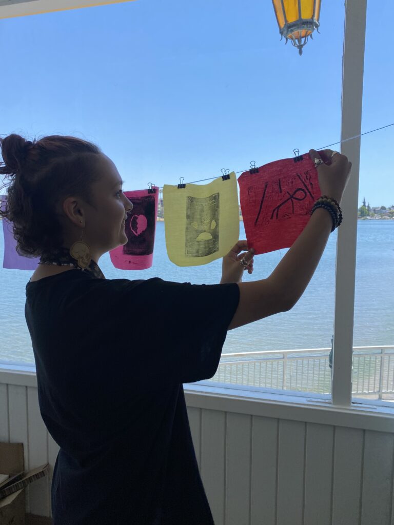 A camper is hanging up a small square piece of red fabric on a clothesline. There is a geometric pattern on the piece of fabric that the camper designed and printed onto it. Lake Merritt is in the backgorund.