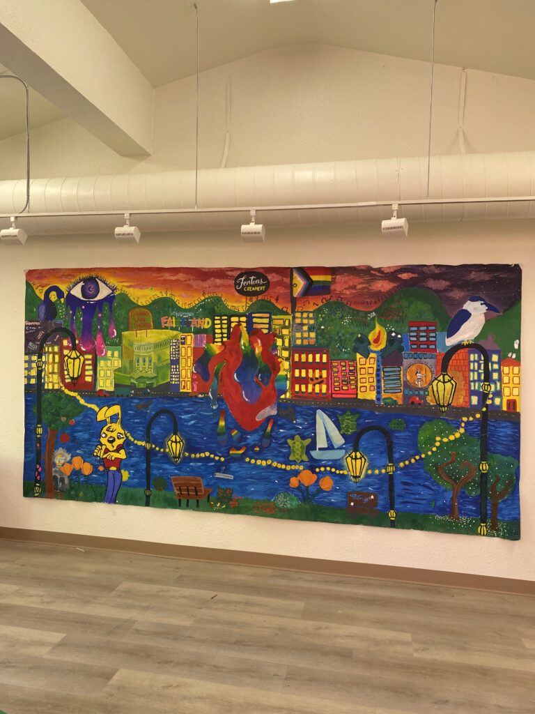 Photo of the mural made in last year's camp. The mural depicts Lake Merritt . In the foreground there is the grassy shoreline, a human-like yellow bunny with clothes on, trees, benches, and the famous String of Lights lamp posts that dot the lake. A night heron is perched on a lamp post. In the background is a sunset and the Oakland cityscape, featuring the Grand Lake Theater, Fairyland, and Fenton's Creamery signs, as well as a LGBT Pride flag and a car on fire. In the center of the mural is a realistic human heart bleeding rainbow colors.